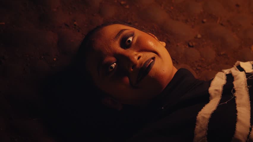 From a top-down view, a woman with theatrical makeup and white eyes lies on the ground in the dark. She slowly forms a sinister, evil smile, looking directly at the camera.