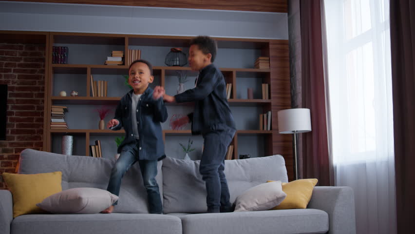 Two little African American kids jumping on couch at living room enjoying relocation moving day in new house funny playful active children sons siblings brothers boys jump on sofa at home playing