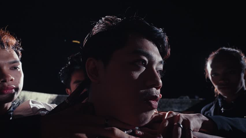 A young man is held in the center of a circle of people in a dark, ritualistic scene. A strong backlight creates a dramatic flare, highlighting the intensity and mystery of the moment.