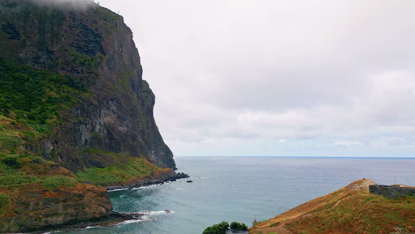 Tall rock standing shoreline aerial view. Waves crashing black stone under cloudy sky. Sheer cliff descending sharply into sea. Green hills extending coastal edge. White clouds hide ridge of mountain.