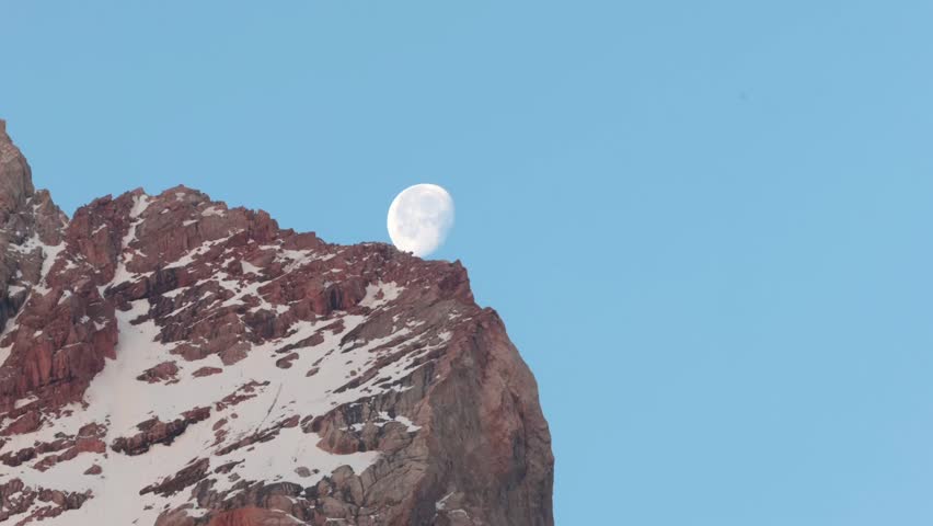 Full moon rises above a snow-capped mountain peak against a clear blue sky. The scene captures the beauty of nature and the tranquility of the night.