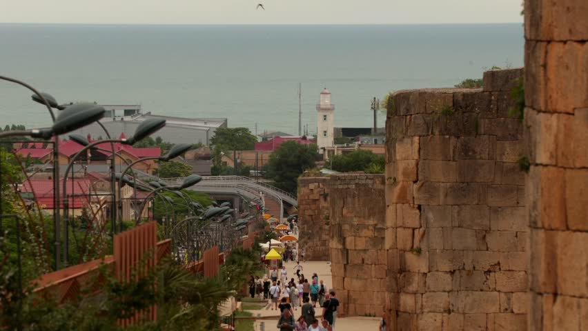 Coastal view with a stone pathway leading down to the beach. People walk along the path, with buildings and a lighthouse visible in the background.