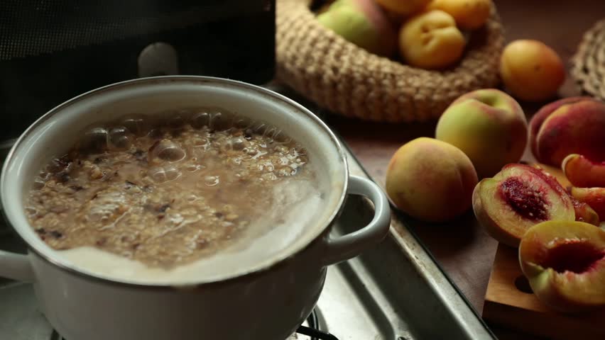 Pot of oatmeal cooking on a stove. Fresh peaches are placed nearby in a basket and on a wooden cutting board. The scene is warm and inviting.