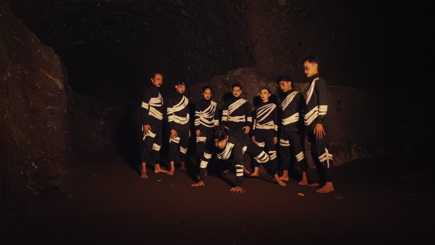 A man in a primal, crawling pose moves toward the camera while his tribe stands behind him with intense expressions. A powerful performance of confrontation or animalistic instinct in a dark cave.