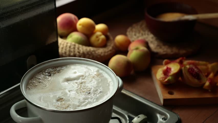Pot of boiling water sits on a stove. Fresh fruits, including peaches and apples, are arranged on a wooden cutting board and in a woven basket nearby.