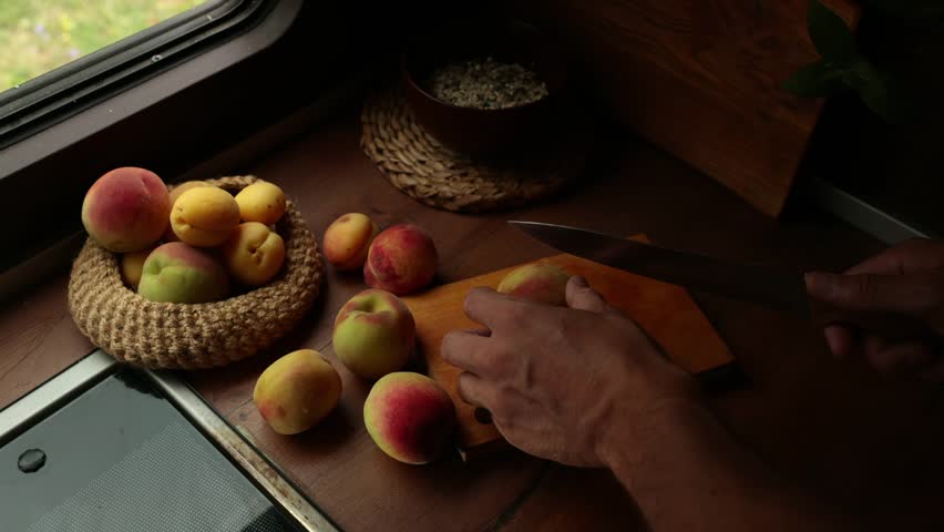 Person with tanned skin slices fresh fruits on a wooden cutting board. Various apples and peaches are arranged nearby in a cozy kitchen setting.