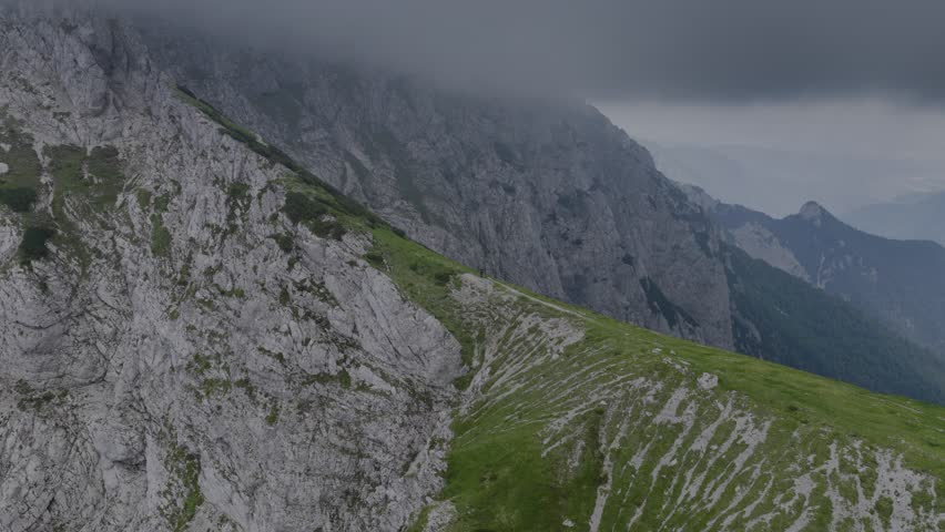 Dramatic Mountain Landscape in Clouds: Stunning view of rocky mountain cliffs partly covered by low clouds and mist, with green slopes below