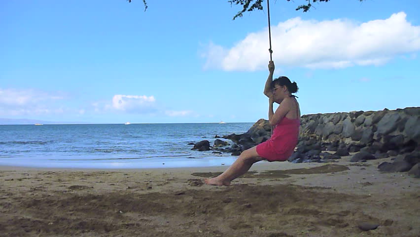 Model released woman sitting on rope swing, enjoying the beach and ocean in