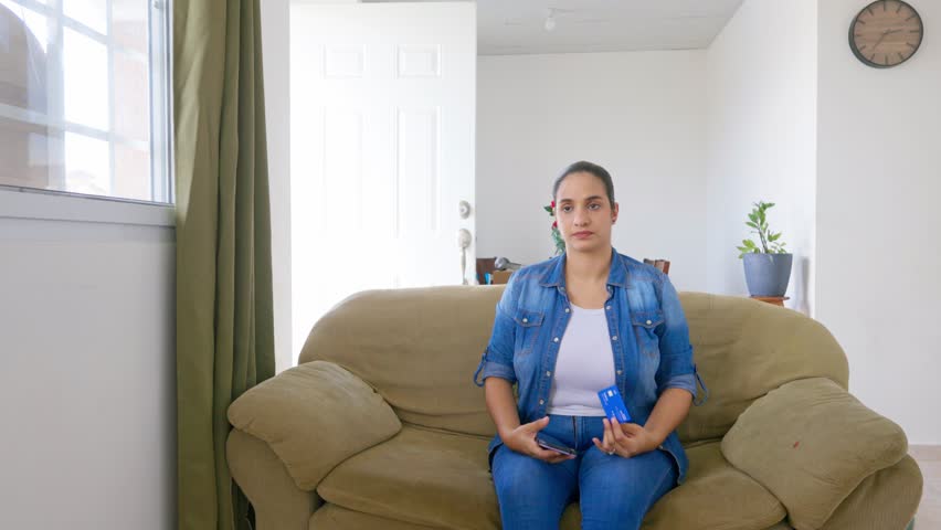 A woman sits on a couch holding a blue card. She is wearing a blue shirt and jeans