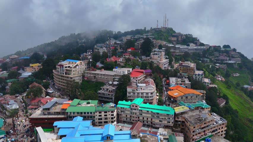 Drone view of a mist-covered hillside town in Murree, Punjab. Packed rooftops, winding roads, and telecom towers rise through forested terrain and active streets.