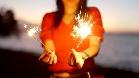 A smiling woman in a red shirt holds sparklers by the sea during sunset. - Powered by Shutterstock - Get 15% off with code: PIKWIZARD15