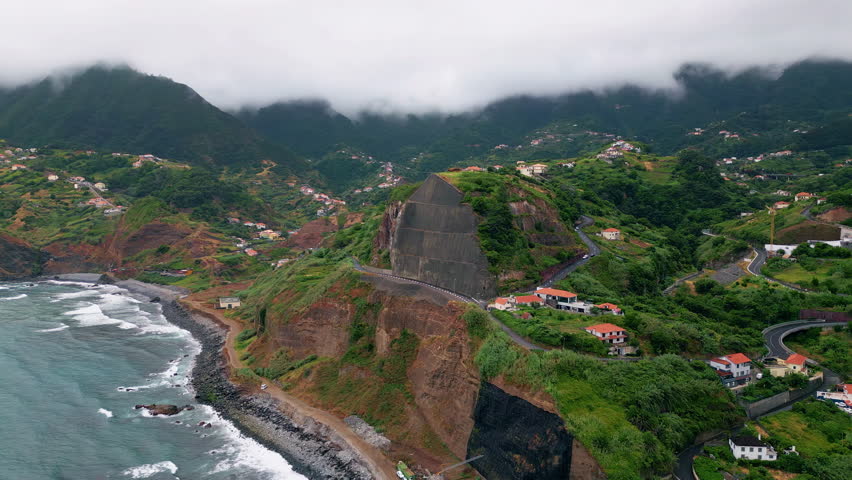 Breathtaking rocky shore drone view. Lush greenery covering mountain hills washed by beautiful ocean waves. Blue sea water splashing foaming on dark stone beach. Picturesque cloudy coastal landscape.