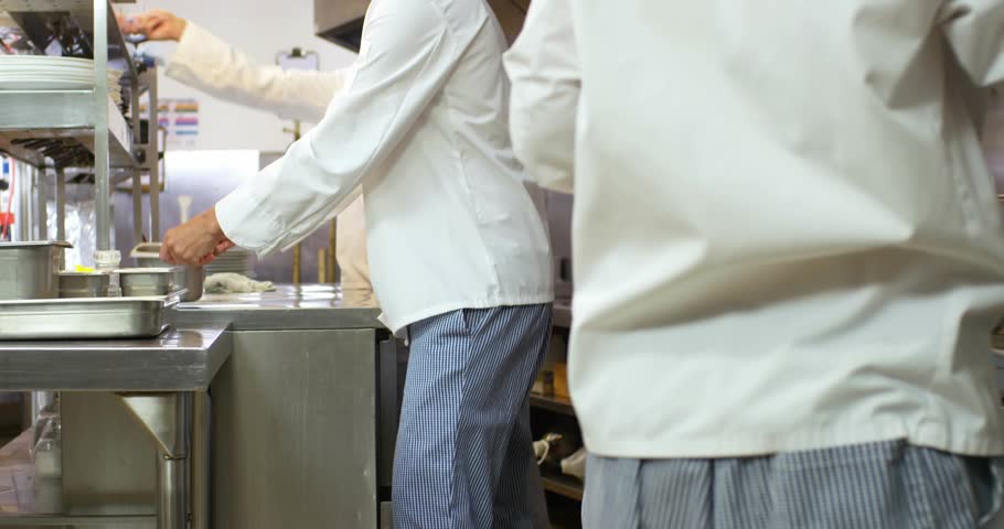 Central chef placing pan triggers chefs loading ovens and stirring batter for plating in kitchen. Professional, teamwork, culinary, stainless steel, busy, seamless, industrial