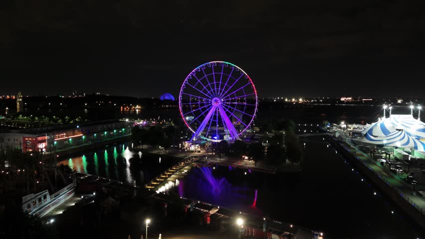 Wide shot of a Ferris wheel illuminated with colorful lights, reflecting in the water at night. Surrounding city lights, tents, and amusement attractions create a lively nighttime atmosphere.