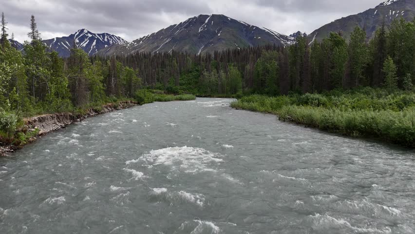 Drone rising above East Fork Chulitna River in Alaska with Trees and Talkeetna Mountains in background, Patches of snow on mountains with Overcast Sky, Thick Dense Forest