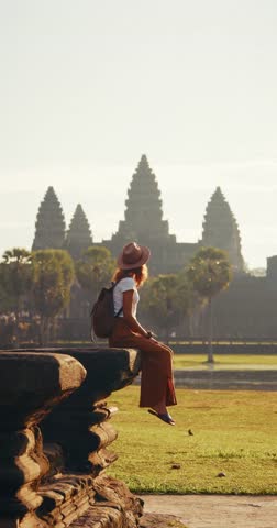 Traveler woman in safari-style hat and backpack sitting casually on stone ledge overlooking Angkor Wat temple in Cambodia. Perfect stock video for lifestyle tourism, cultural promo, and travel ads.