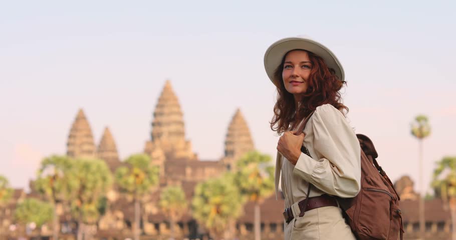 Traveler woman in stylish outfit, hat and backpack looks to the side then turns toward Angkor Wat temple in Cambodia. Concept of lifestyle, adventure, tourism, promo, advertisement and cultural travel