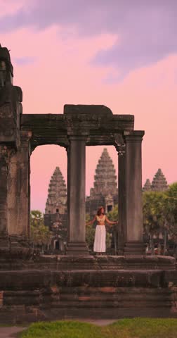 Young woman in boho outfit exploring Angkor Wat temple in Cambodia at pink sunset. Concept of travel, lifestyle, adventure, cultural tourism, promo, advertisement and exotic destinations.