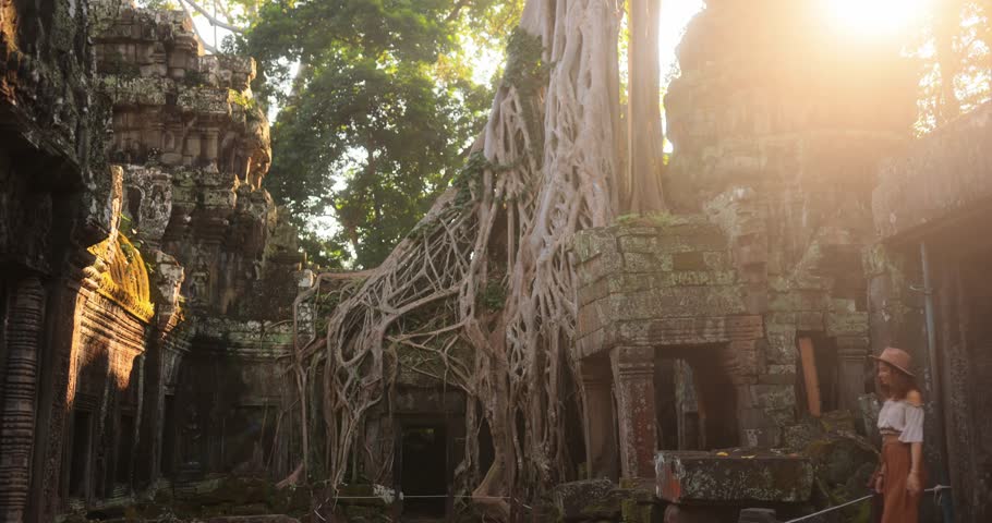 A female traveler in a hat passes by and enters Ta Prohm temple at Angkor Wat, Cambodia, as sun rays shine through jungle trees. Perfect adventure tourism and cultural heritage promo footage.