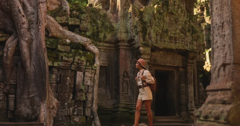 Young woman with hat and backpack explores the famous Ta Prohm Temple where massive tree roots cover ancient Khmer ruins. Perfect stock video for travel blogs, cultural documentaries, and tourism