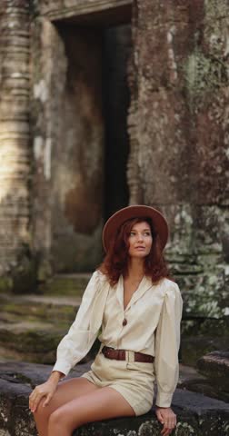 Elegant woman with red hair and hat sitting at Ta Som temple in Angkor Wat, Cambodia. Conceptual travel lifestyle stock video for tourism, culture, fashion promo and advertisement campaigns.