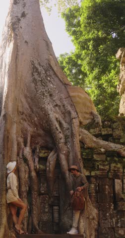 Man and woman explore Ta Prohm temple at Angkor Wat, Cambodia, walking past massive tree roots covering ancient ruins. High quality travel footage for documentaries, tourism and cultural promos.