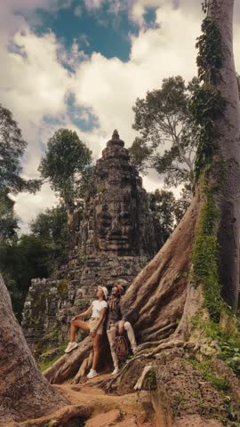 Young couple in casual travel outfits relaxing by giant tree roots near Victory Gate, Bayon Temple, Angkor Thom, Cambodia. High quality lifestyle and tourism stock footage for promo and adventure ads.
