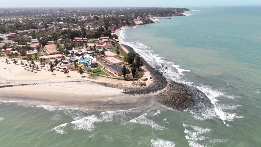 Drone footage of Banjul, Gambia, with coastal houses and palm trees near the turquoise Atlantic waters along the shoreline.
