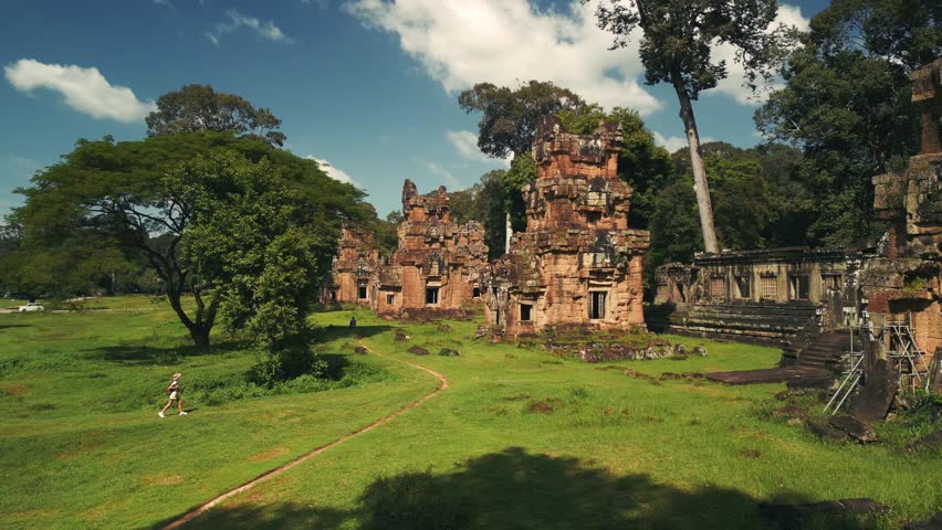 Smooth drone shot tracking a woman tourist in safari clothes exploring ancient Suor Proat temple near Bayon, Angkor Thom, Cambodia. Concept of adventure travel, heritage tourism, and cultural promo.