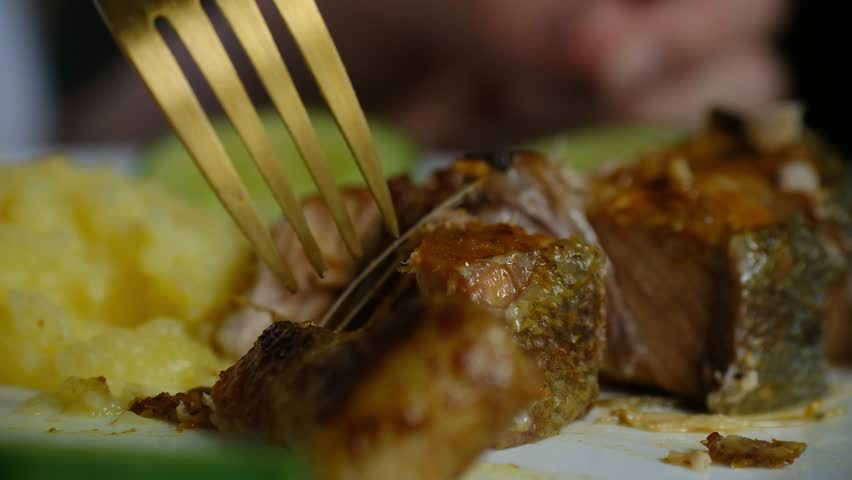 Close up person use golden fork to break off piece of juicy baked salmon, served with mashed potatoes, fresh cucumber slices on white plate during gourmet meal. Golden fork flaking delicious salmon.