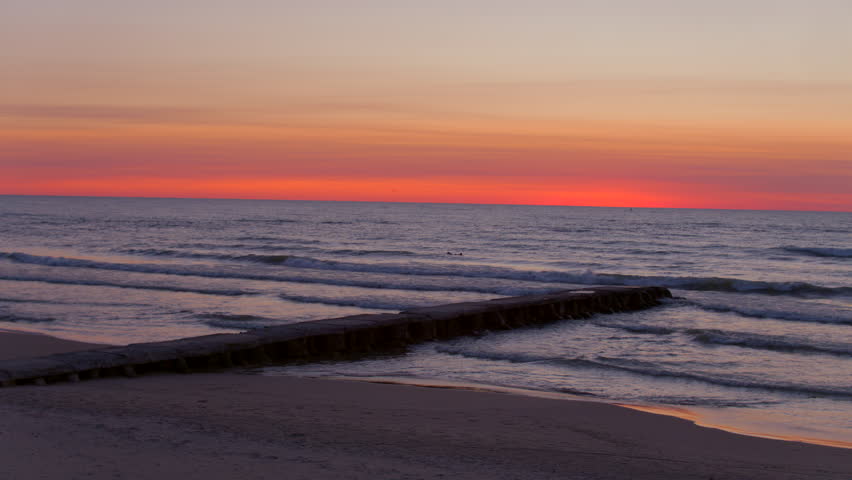 Drone aerial at dawn over Lake Michigan trucking right along the shoreline pier as a surfer paddles through gentle waves beneath a glowing morning horizon.