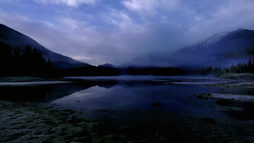 A serene lake reflects the misty mountains under a cloudy twilight sky
