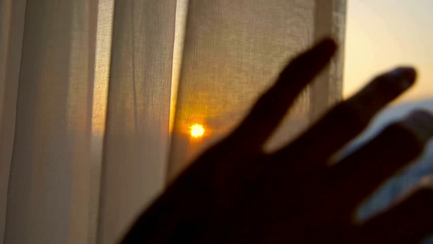 A silhouetted hand opens a window curtain to reveal a beautiful golden sunrise over the city. A concept of hope and a new day.