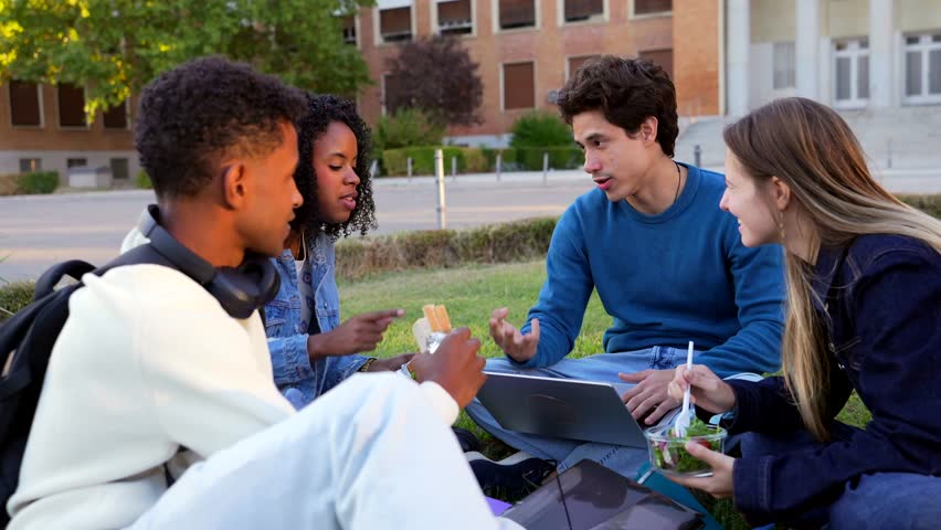 Group of four multiracial college students sitting on the grass, studying with a laptop, and eating lunch together, happily chatting and collaborating on a university campus lawn