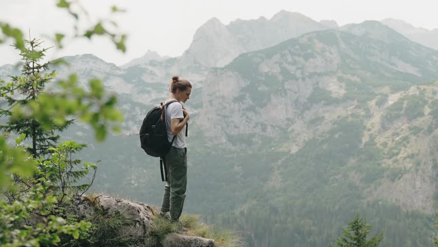 A young woman traveler with a hiking backpack stands on the edge of a cliff, admiring the panoramic view of the mountains
