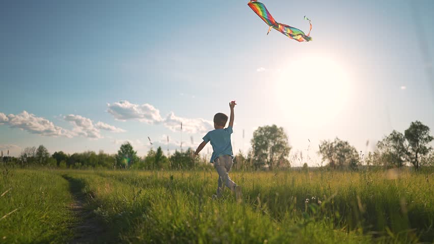 Boy running outdoors under summer sun. Child lifts arm flying kite. Happy carefree kid enjoys freedom. Boy laughs running free. Outdoors makes child happy. Flying kite brings carefree summer freedom.