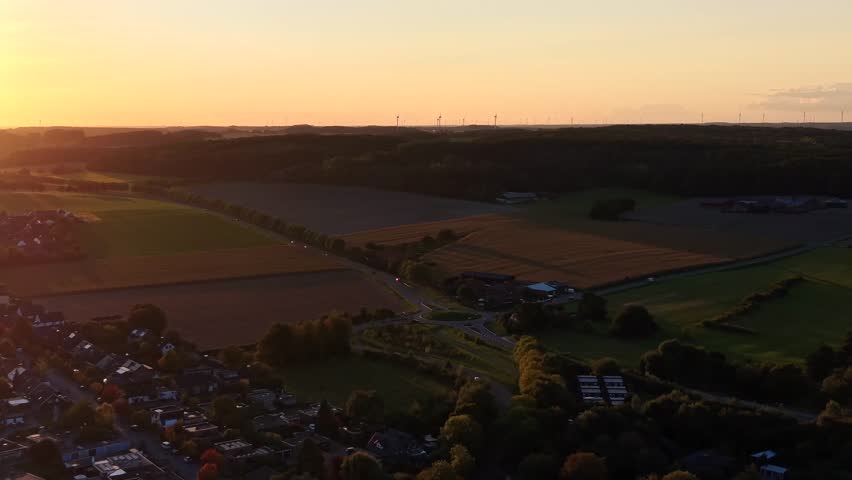 Golden sunset in small American town with traffic on roundabout. Aerial approaching wide shot. Houses and homes of suburb neighborhood near farm fields and green hills in late summertime.