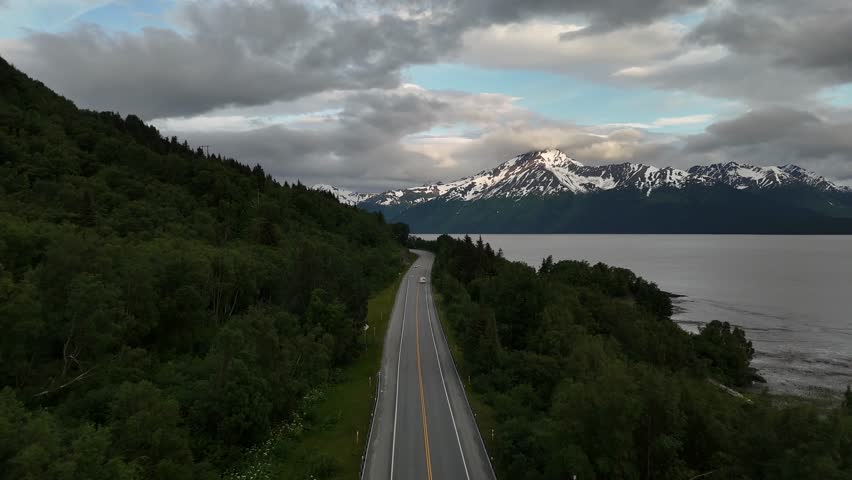 Aerial View, Car Driving Down Road surrounded by trees, Seward Highway in Alaska along Turnagain Arm, Snowcapped Mountains in Background Toward Late Evening in Mid June