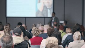 Crowd of professionals sits in rows facing large screen as speaker presents data and slides in bright contemporary conference space. - Powered by Shutterstock - Get 15% off with code: PIKWIZARD15