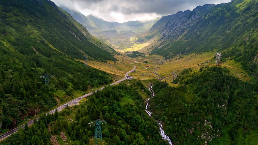 Spectacular green mountains from drone footage. A highway and narrow river pass among the rocks. Grey rocks cover the rocks at backdrop. Romania aerial view.