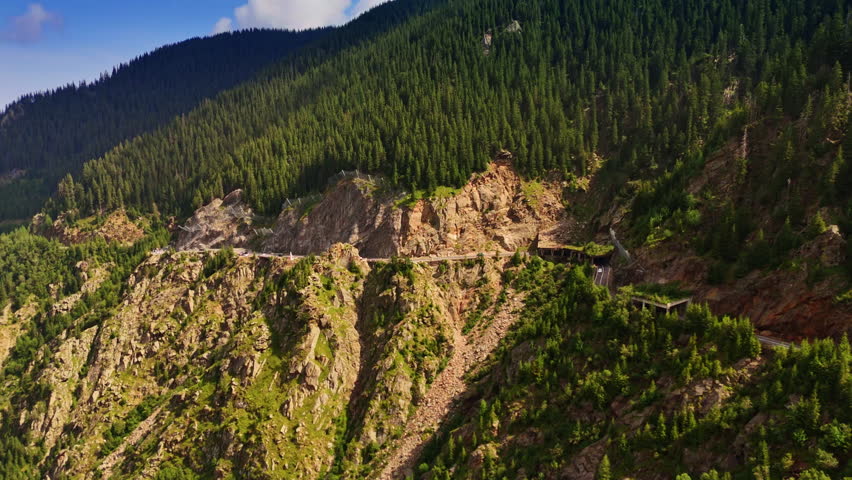 Approaching a spectacular highway situated on the mountain slope. Pine tree woods cover the cliff rocks. Romania from drone footage.