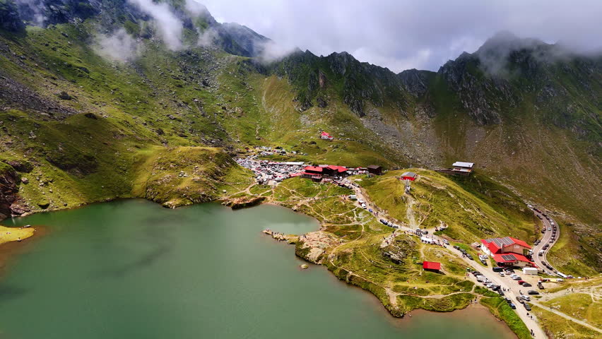 Flying over the green mountain lake with touristic infrastructure nearby. Lots of people and cars are near the waterscape. Breath-taking scenery of Romania.