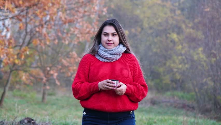 Close up of a woman in red warm knitted sweater drinking hot tea from the thermos mug walking in the autumn forest