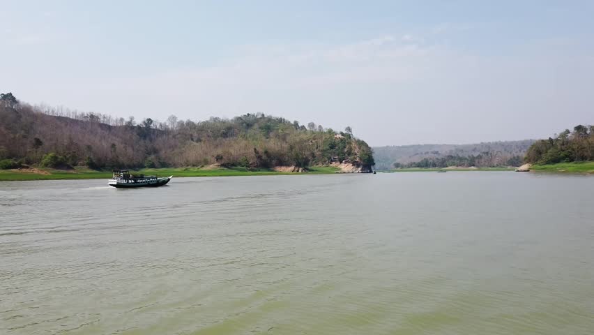 A traditional boat glides across a wide, calm river, flanked by verdant green banks and tree-covered hills under a bright, clear sky.
