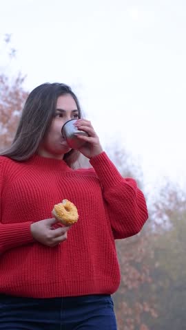Close up of a woman taking a bite of glazed donut, walking in autumn forest wearing red sweater and scarf