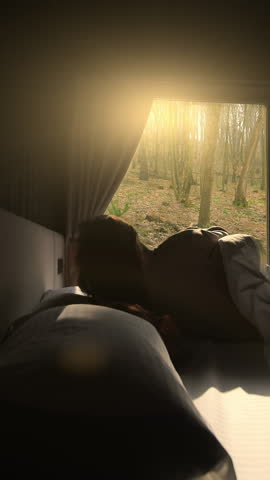 A person sits on a bed in a dark room looking out of a window as the morning sun rises over a forest. The scene captures the calm and serene atmosphere of waking up in nature.