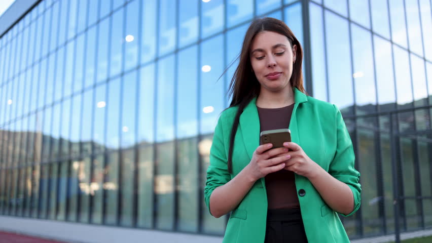Cheerful young businesswoman strolling by a sleek glass office building, with a smile on her face as she types and texts on her smartphone