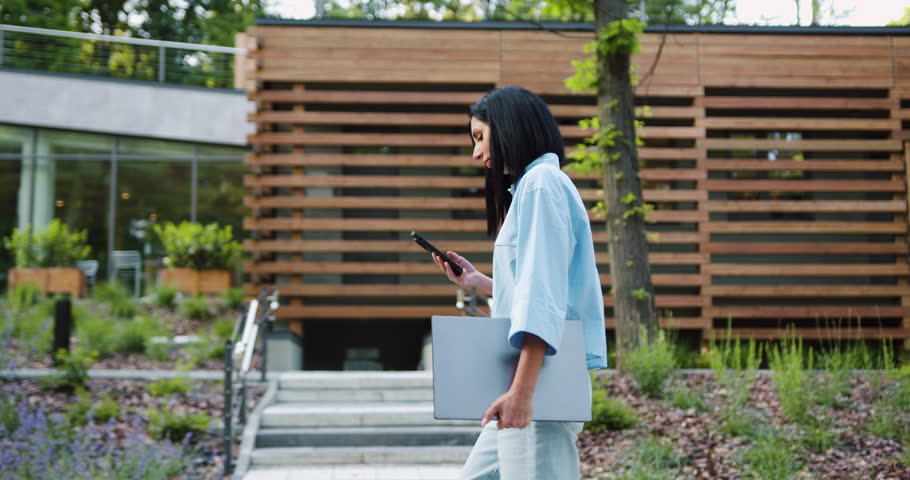 Side view of young Caucasian business woman in casual outfit use smartphone and holding laptop in hand while walking at front of modern building at city park. Business and people concepts