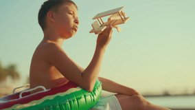 Child boy sitting alone on sandy beach on sea coast and playing with toy plane. Cinematic slow motion shot, bored kid viewing wooden aircraft model and dreaming of flight and traveling to new location - Powered by Shutterstock - Get 15% off with code: PIKWIZARD15