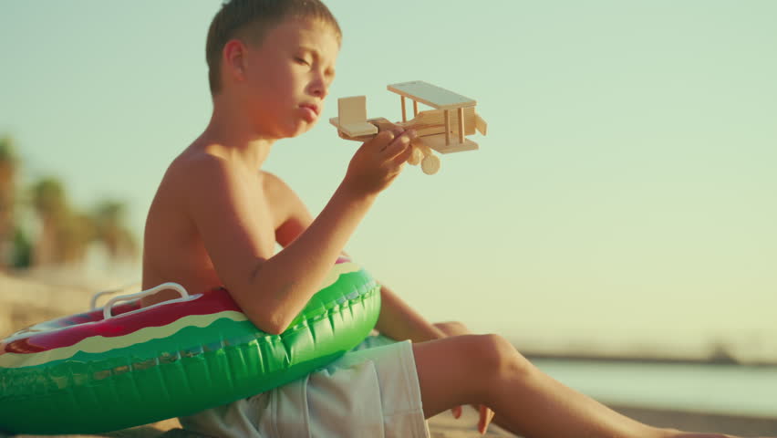 Little boy dreaming of aviation, imaging how flying by plane, future pilot. Caucasian child playing with toy airplane in summer, resting on sandy beach and warming on sun in morning, slow motion shot